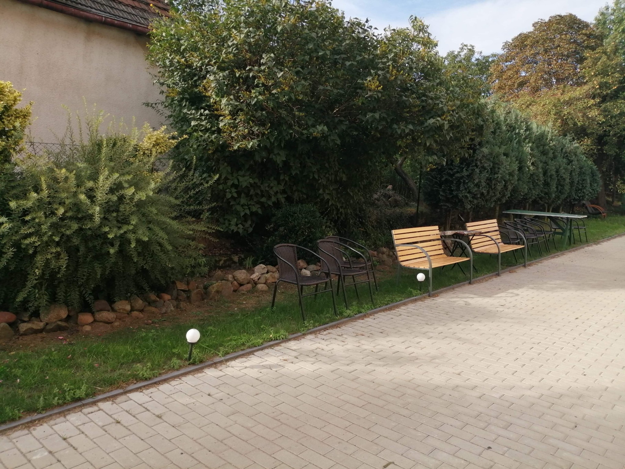 Zewnętrzny teren relaksacyjny z drewnianymi ławkami i krzesłami z wikliny, otoczony zielenią i oświetlony kulistymi lampami, wyłożony kostką brukową.