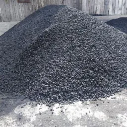 Góra czarnego, błyszczącego paliwa stałego na placu składowym, widoczny drewniany płot i tabliczka informacyjna.