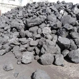 Góra czarnego węgla kamiennego o nieregularnych kształtach, z widocznymi śladami pyłu węglowego na podłożu i ścianie w tle.