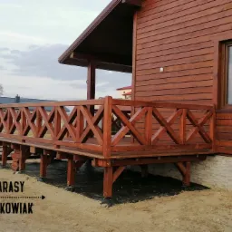 Drewniany taras przydomowy z balustradą w kratkę, widoczny fragment elewacji domu z drewnianą boazerią i oknem, teren wokół tarasu jeszcze niezagospodarowany.