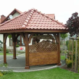 Drewniana altana z czerwonym, ceramicznym dachem w ogrodzie, z widoczną kratką na jednej ze ścian i ozdobnymi łukami podtrzymującymi dach.