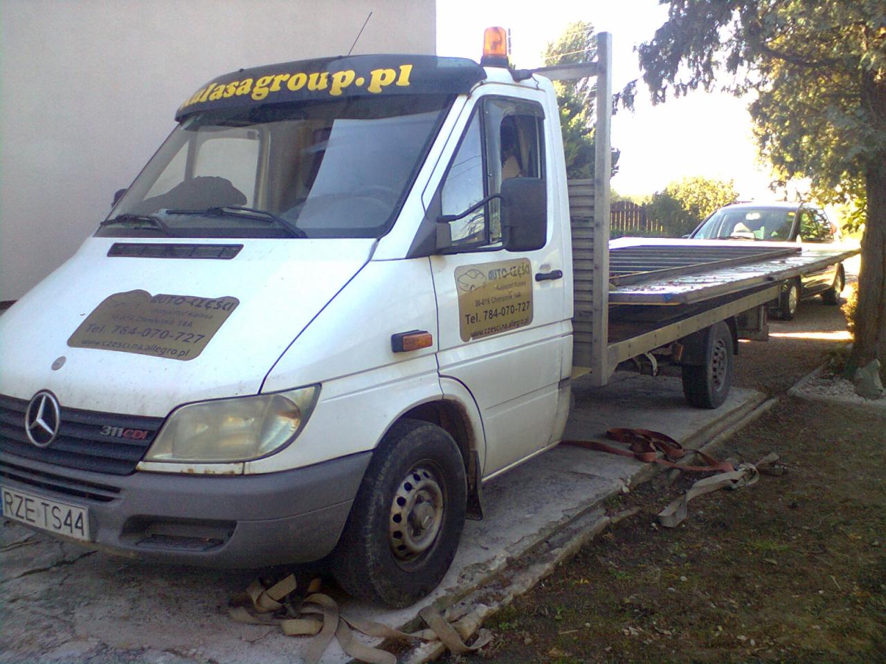 Biała laweta Mercedes-Benz 311 CDI z żółtą lampą ostrzegawczą na dachu, częściowo załadowana samochodem osobowym, na betonowym podjeździe przed budynkiem i drzewem, pasy zabezpieczające leżą na ziemi.