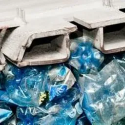 Strumień zgniecionych, niebieskich butelek PET w sortowni odpadów, pod metalową konstrukcją podajnika.