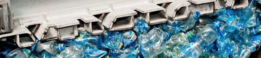 Strumień zgniecionych, niebieskich butelek PET w sortowni odpadów, pod metalową konstrukcją podajnika.