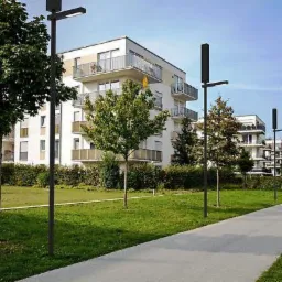 Ścieżka spacerowa w parku oświetlona nowoczesnymi, czarnymi latarniami LED na tle białych budynków mieszkalnych z balkonami.