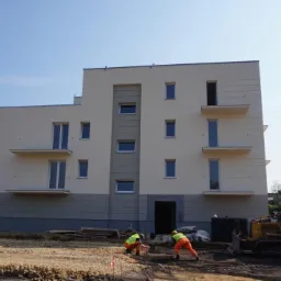 Prace ziemne przy nowo wybudowanym, trzypiętrowym budynku mieszkalnym z balkonami; dwóch robotników w odblaskowych kamizelkach i kaskach pracuje na placu budowy, w tle żółta spycharka CAT 936.