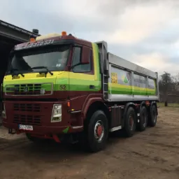 Ciężarówka wywrotka Terberg z numerem 52, z zielono-żółto-brązowym malowaniem, zaparkowana na placu obok szarego budynku z otwartą bramą, widoczna tablica rejestracyjna GST-76881 i numer telefonu...
