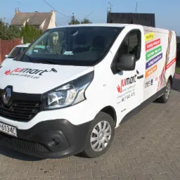 Biały samochód dostawczy Renault z logo Jumart i informacjami kontaktowymi firmy specjalizującej się w instalacjach elektrycznych, monitoringu i napędach bram, zaparkowany na brukowanej ulicy przed...