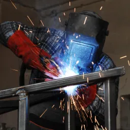 Spawacz w masce ochronnej i czerwonych rękawicach łączy metalowe elementy konstrukcji, widoczne iskry i dym.