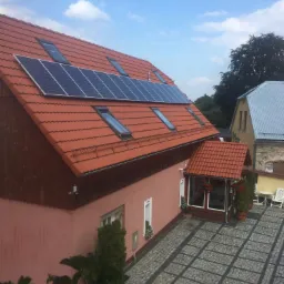 Czerwony dach skośny z panelami słonecznymi i oknami dachowymi, widok z góry na budynek z różową elewacją i brukowanym podjazdem.