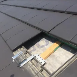 Fragment dachu z ciemnej dach&oacute;wki ceramicznej w trakcie montażu, widoczne elementy mocujące i fragment membrany dachowej, drewniana łata z zielonym znacznikiem.