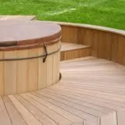 Drewniany taras z okrągłą zabudową wanny jacuzzi, widoczne schodki i zakrzywiona balustrada, trawnik w tle.