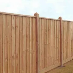 Drewniane ogrodzenie panelowe z pionowych desek, zakończone słupkami z piramidalnymi zwieńczeniami, na tle trawnika i oddalonego krajobrazu.