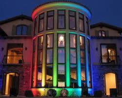 Nowoczesna, przeszklona, cylindryczna konstrukcja balkonu z wielokolorowym oświetleniem LED, wkomponowana w fasadę budynku. Widoczne elementy wystroju wnętrza przez duże okna.