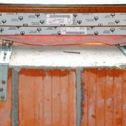 OPTIMA Sebastian Dudek - Fragment montażu okna z widocznymi metalowymi kotwami mocującymi do muru z cegły ceramicznej, uszczelnienie folią wiatroizolacyjną, widoczny fragment ramy okiennej z naklejkami producenta.