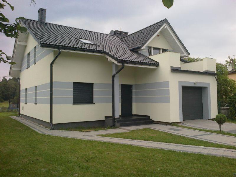 Dwukondygnacyjny dom z szarą stolarką okienną i poziomymi pasami na elewacji, otoczony trawnikiem i brukowanym podjazdem.