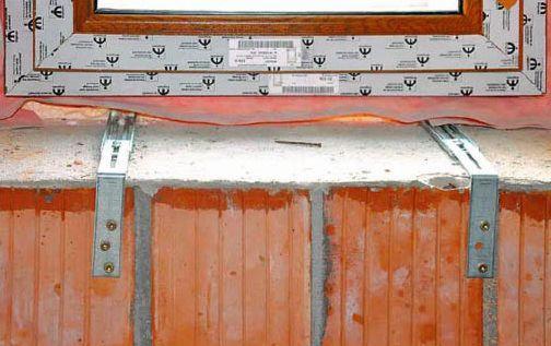 Fragment montażu okna z widocznymi metalowymi kotwami mocującymi do muru z cegły ceramicznej, uszczelnienie folią wiatroizolacyjną, widoczny fragment ramy okiennej z naklejkami producenta.