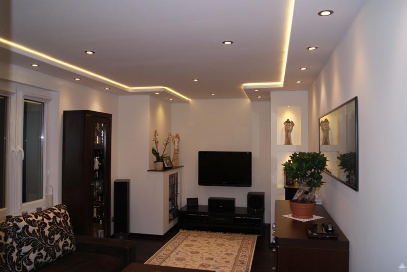 Nowoczesny salon z podwieszanym sufitem z oświetleniem LED, punktowe lampy, białe ściany, telewizor, szafka RTV, dywan, dekoracje, roślina w doniczce i ciemne meble.