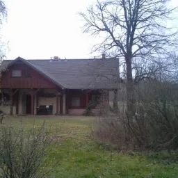 Dom jednorodzinny z ciemną, spadzistą dachówką i drewnianą elewacją, widziany z perspektywy ogrodu w pochmurny dzień. Widoczne drzewa bez liści.