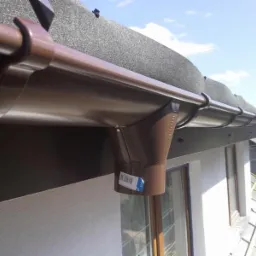 Brązowy system rynnowy zamontowany na dachu budynku, widoczny z bliska z naklejką na rurze spustowej. Dach pokryty szarą papą. Fragment okna i ściany budynku.