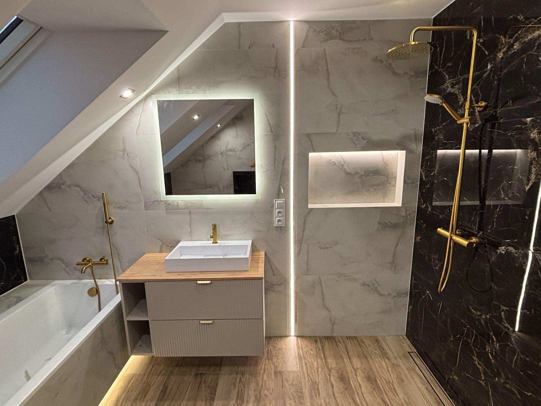 Nowoczesna łazienka na poddaszu z marmurowymi płytkami, złotą armaturą i podświetlanym lustrem, drewnopodobna podłoga, widoczna wanna i kabina prysznicowa.