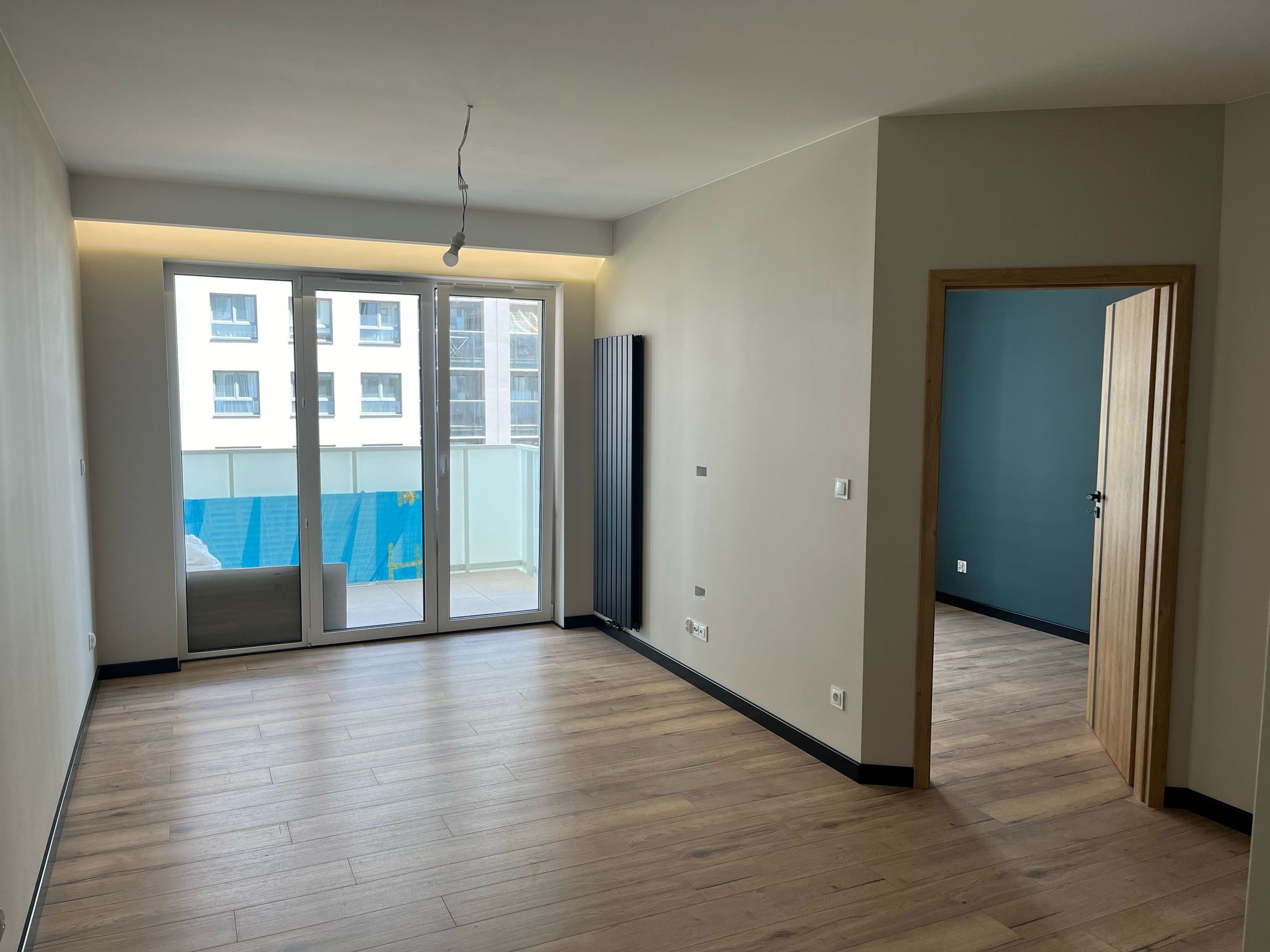 Pusty pokój z drewnopodobną podłogą, dużym oknem balkonowym z widokiem na budynek i balkon, oraz otwartymi drzwiami do pokoju z pomalowaną na niebiesko ścianą.