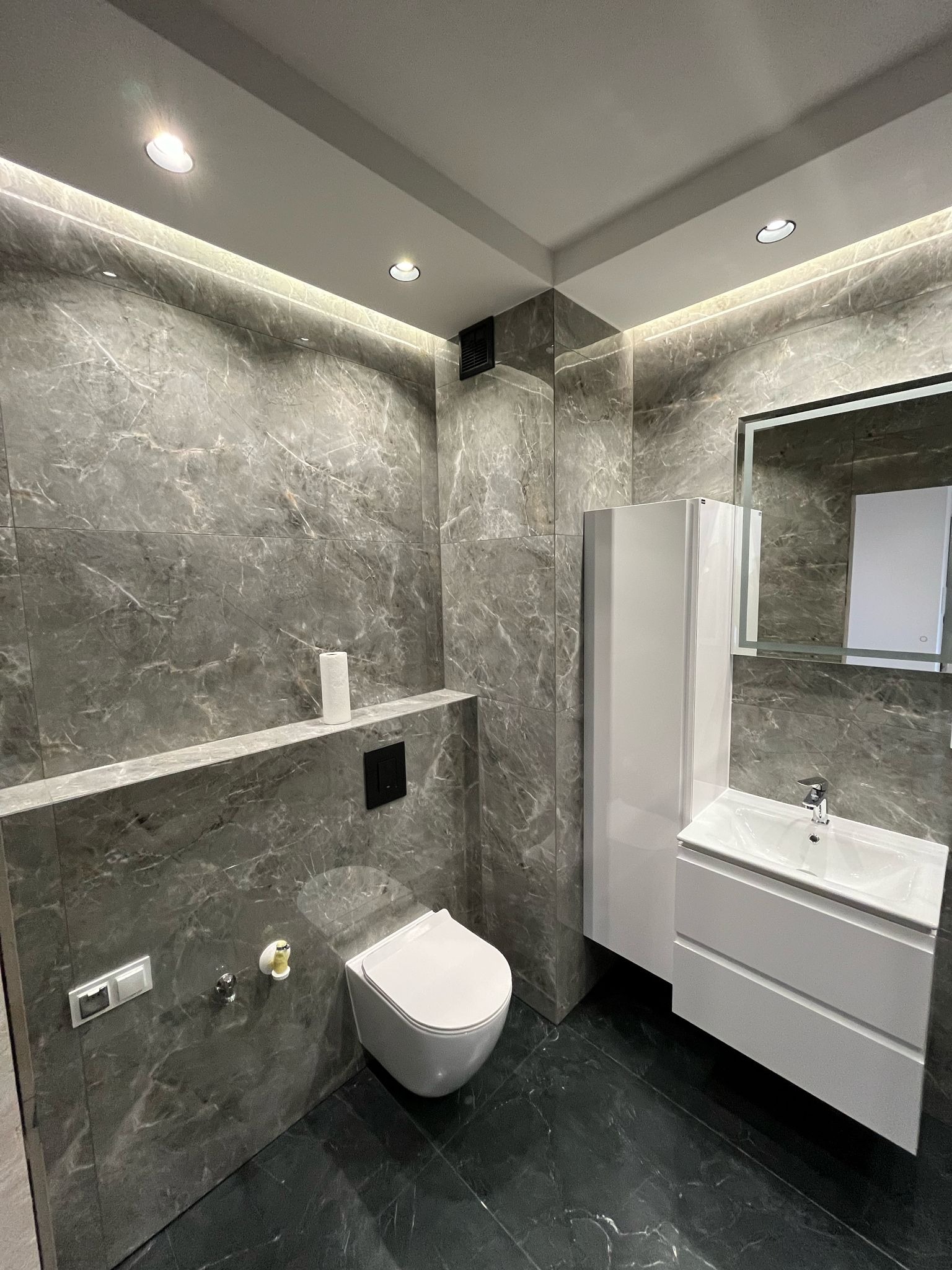 Nowoczesna łazienka z szarymi marmurkowymi płytkami, podświetleniem LED, wiszącą toaletą i białą szafką z umywalką.