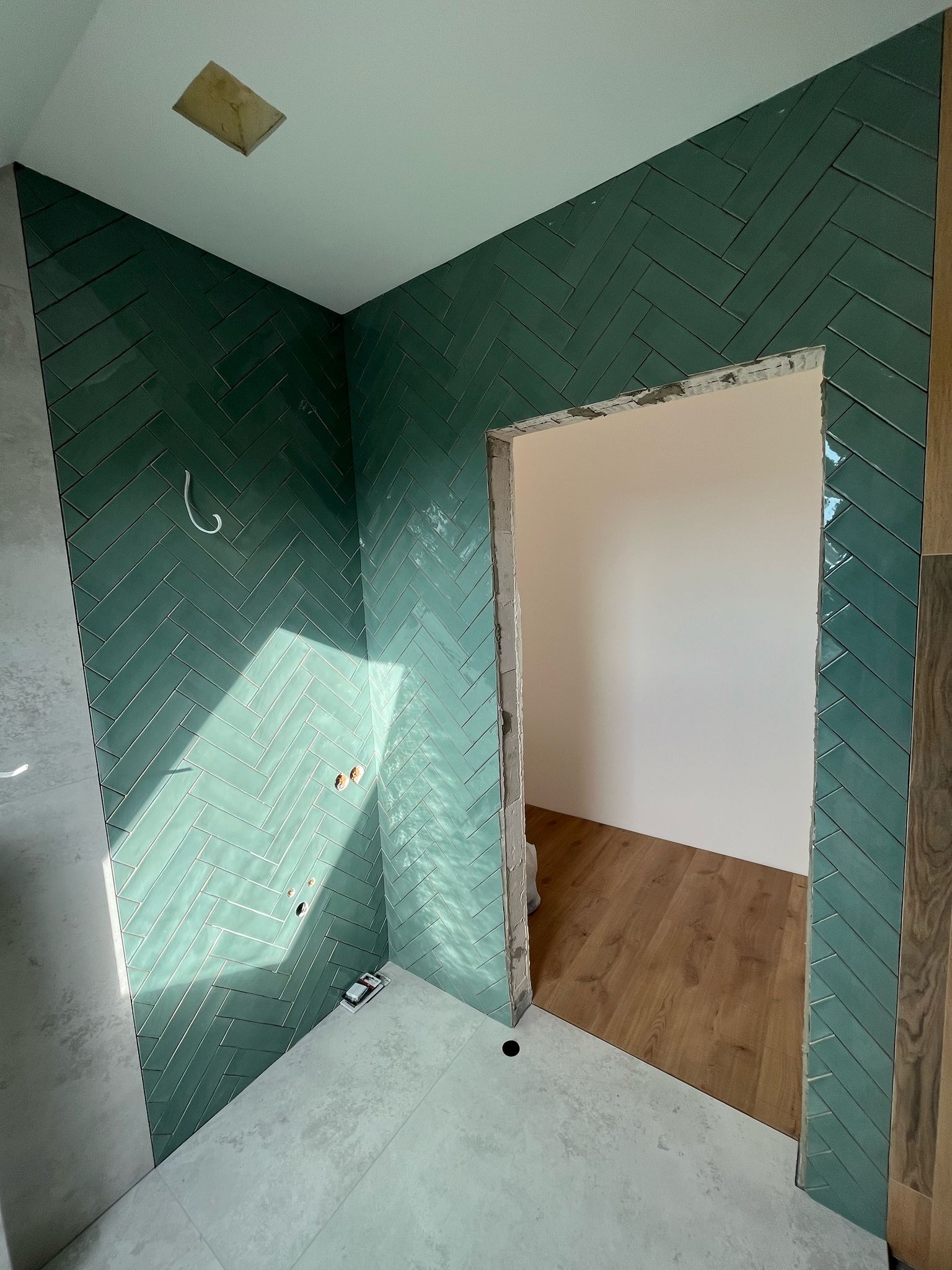 Łazienka w trakcie wykańczania: ściany wyłożone zielonymi, błyszczącymi płytkami ułożonymi w jodełkę, surowy otwór drzwiowy z widocznym klejem, podłoga częściowo wyłożona szarymi płytkami,...