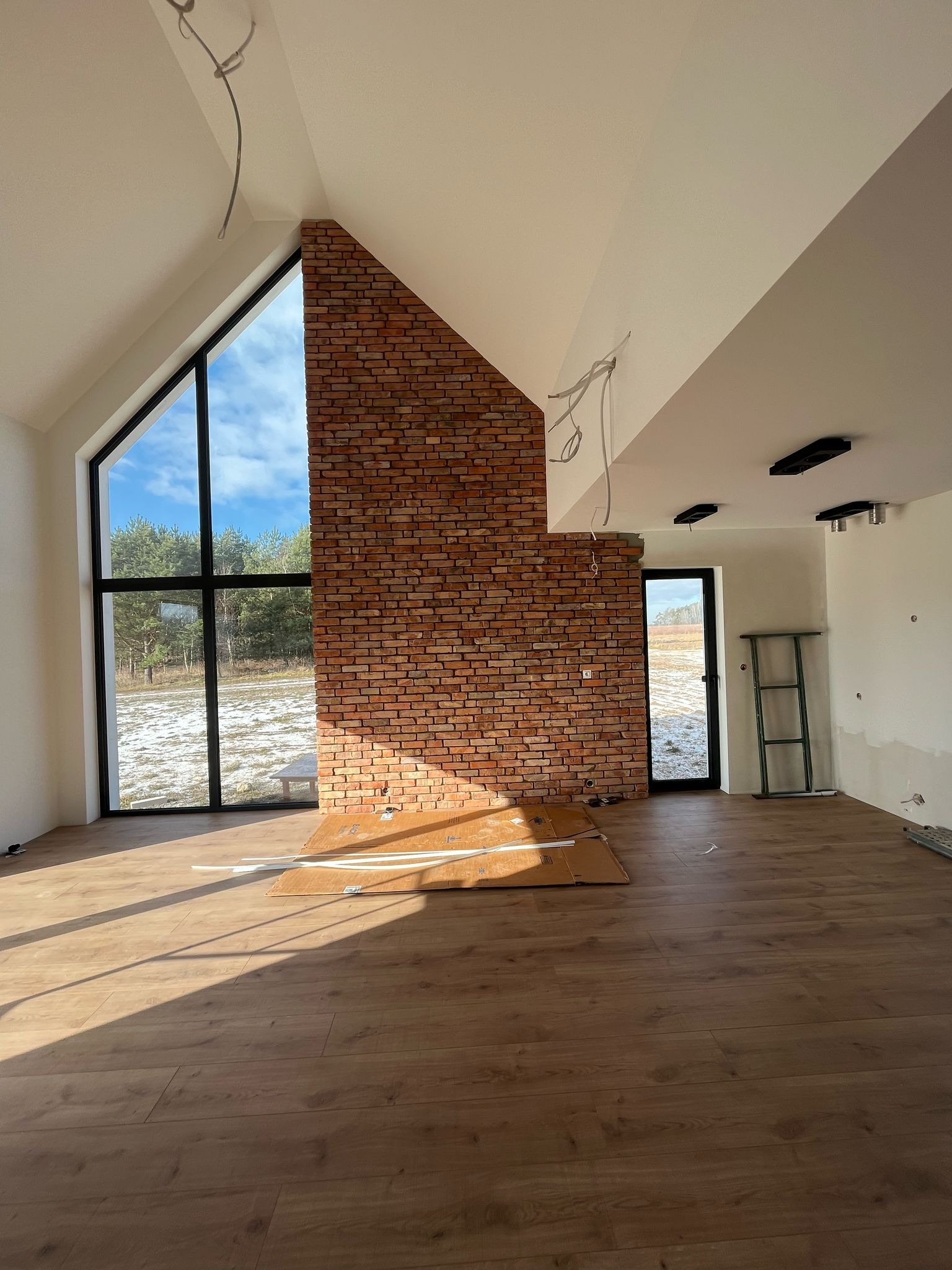 Wnętrze w trakcie wykańczania: drewnopodobne panele podłogowe, ściana z czerwonej cegły, duże okno z widokiem na ośnieżone pole, widoczne kable wystające z sufitu.