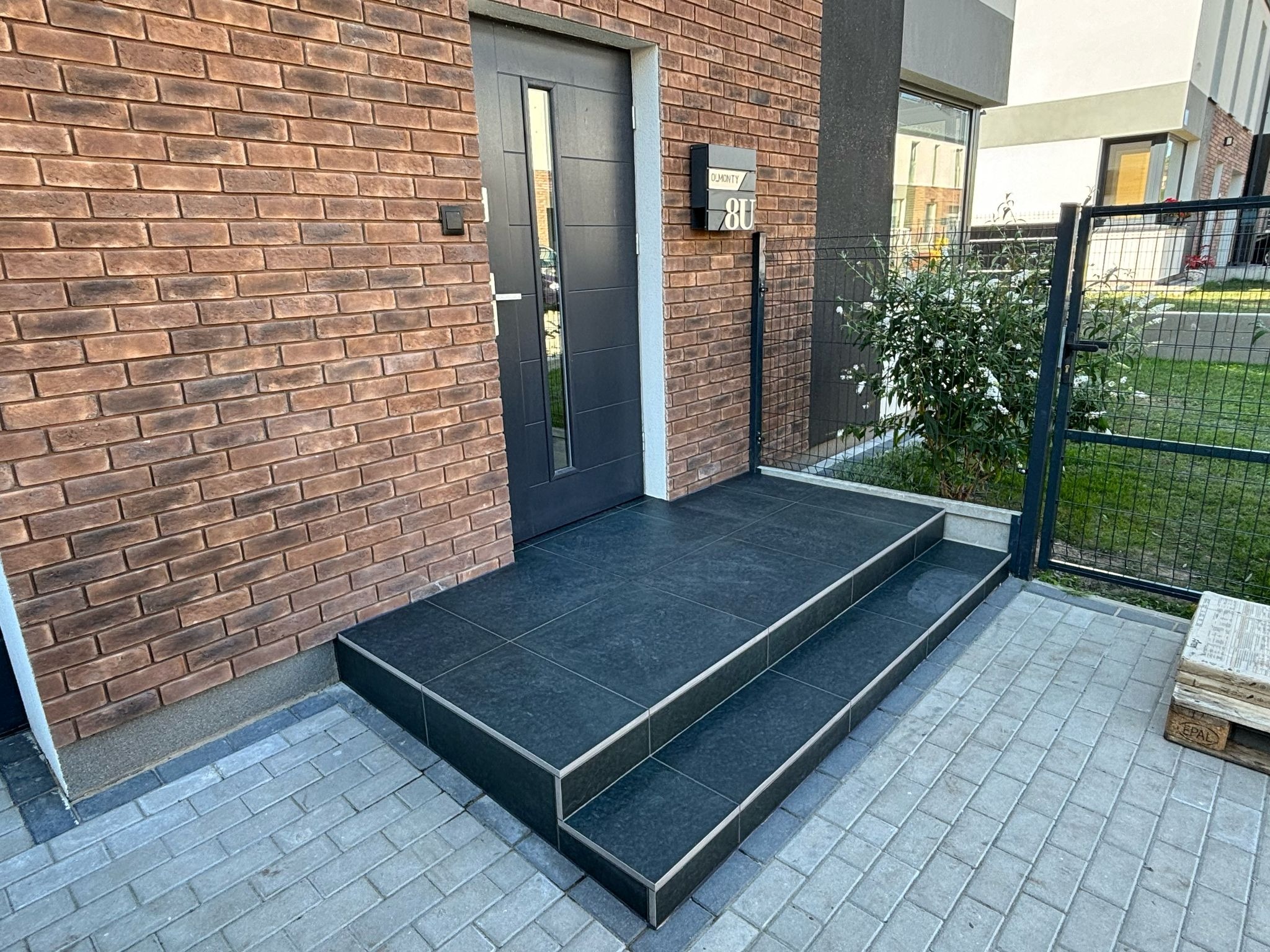 Nowoczesne wejście do domu z dwustopniowymi schodami wyłożonymi ciemnymi płytkami z metalowymi listwami wykończeniowymi, ceglana elewacja i czarne drzwi.