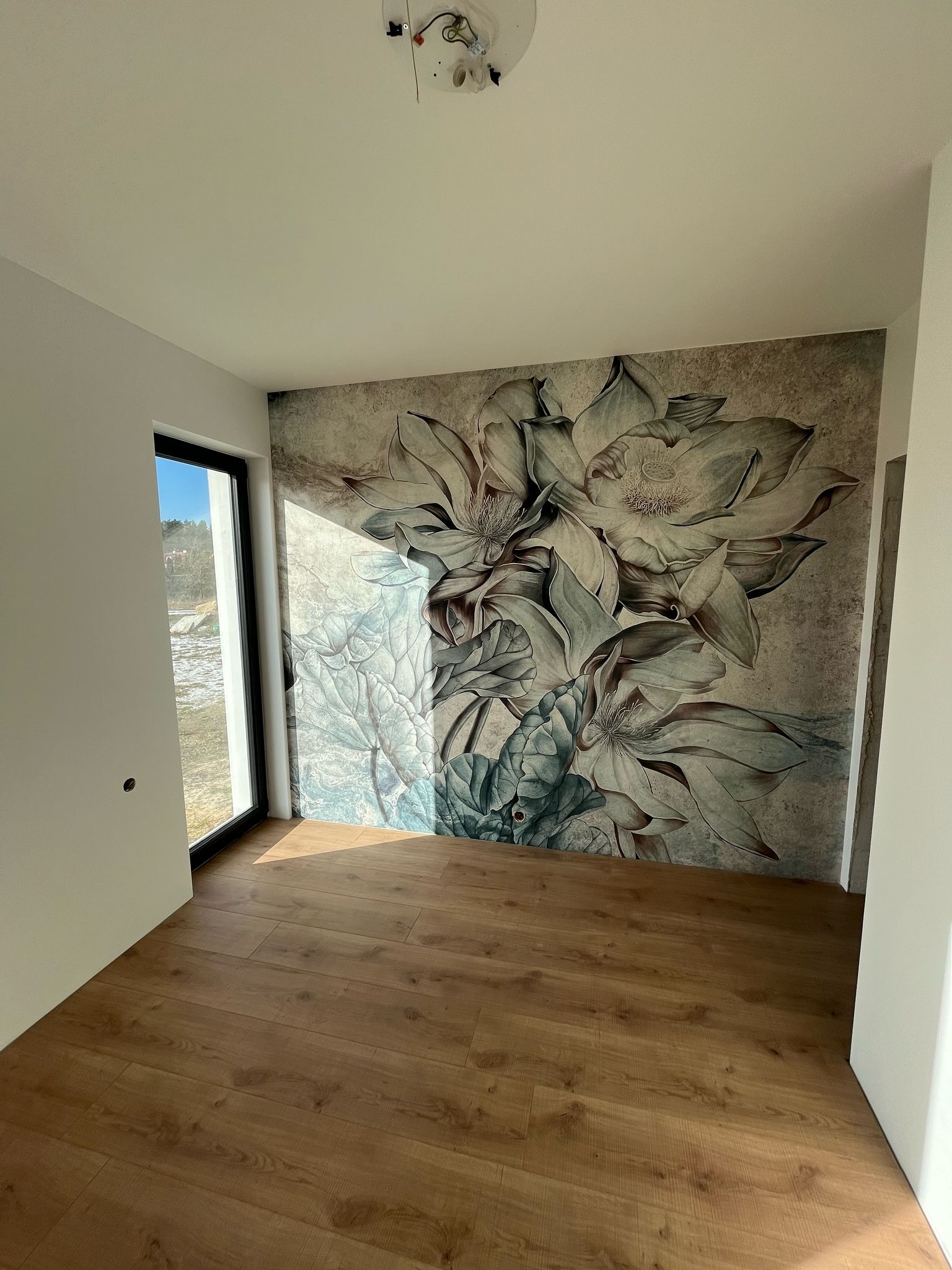 Jasne panele podłogowe w pokoju z fototapetą przedstawiającą duże kwiaty lotosu na ścianie, widok z okna na zewnątrz.