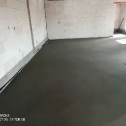 Świeżo wylana, gładka, szara posadzka w surowym wnętrzu budynku z białych bloczków, widoczne folie przy ścianach i fragment niebieskiej rurki w tle.