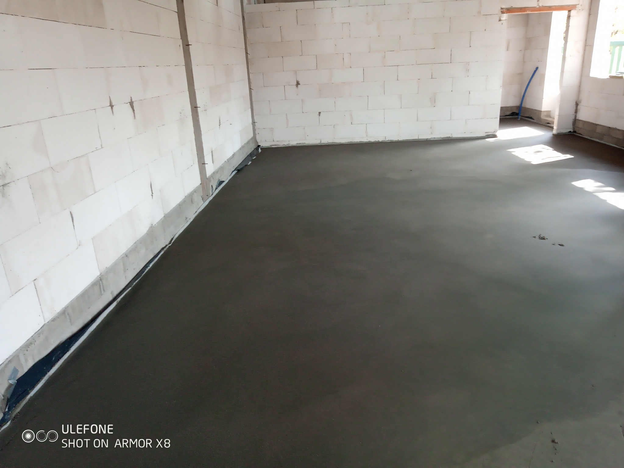 Świeżo wylana, gładka, szara posadzka w surowym wnętrzu budynku z białych bloczków, widoczne folie przy ścianach i fragment niebieskiej rurki w tle.