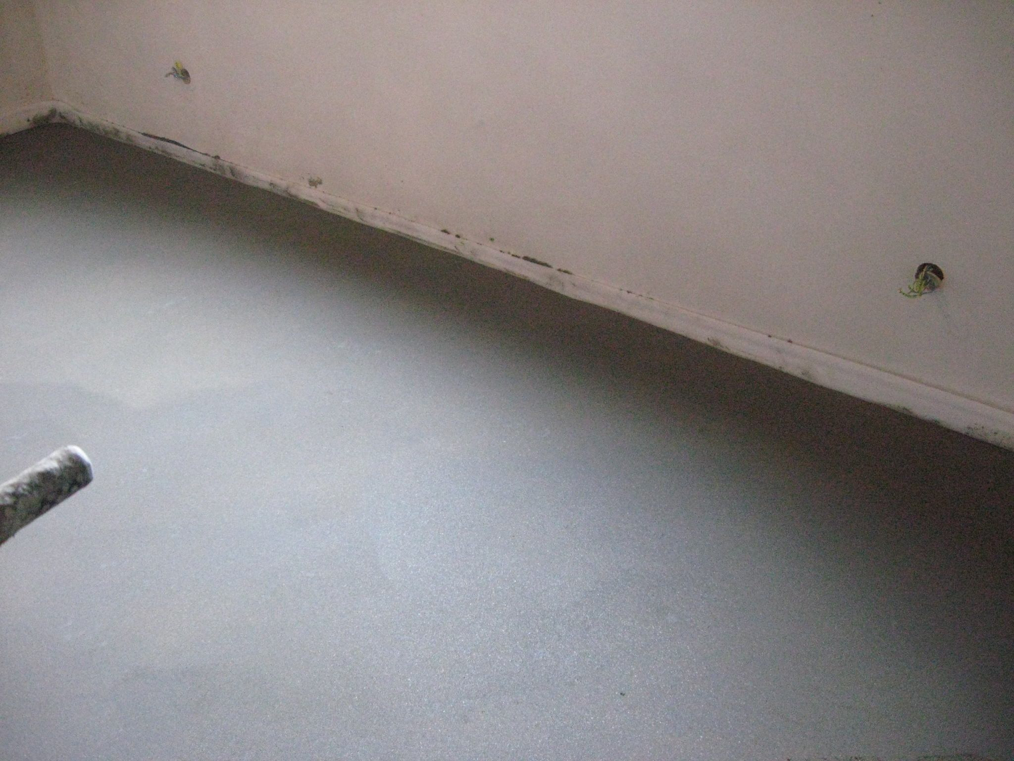 Świeżo wylana, gładka posadzka w pomieszczeniu, widoczne przewody elektryczne wystające ze ściany oraz fragment narzędzia do rozprowadzania masy.