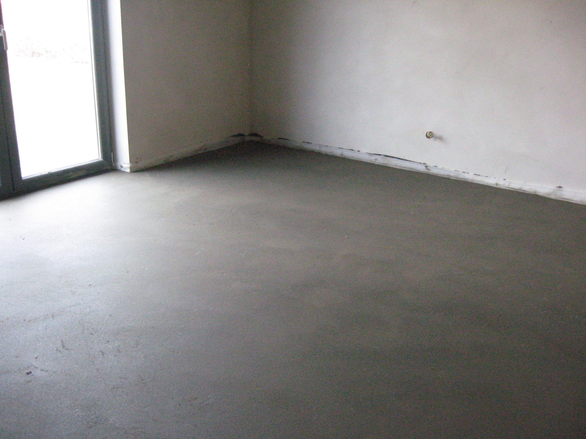 Świeżo wylana, gładka posadzka jastrychowa w pustym pomieszczeniu z dużym oknem balkonowym, widoczne ślady po narzędziach i listwa dylatacyjna przy ścianie.