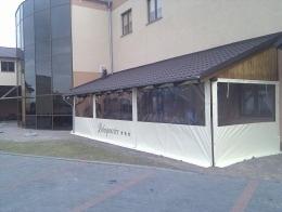Zadaszony ogródek restauracyjny z białymi ścianami bocznymi i brązowym dachem, przylegający do budynku z przeszkloną fasadą.