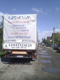 Ciężarówka z plandeką reklamową firmy ArtKlim, specjalizującej się w klimatyzacji. Widoczny numer telefonu i adres strony internetowej na białej plandece. Ulica z kałużami.