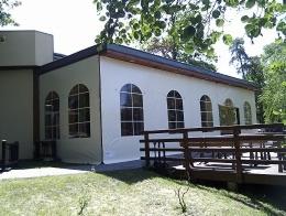 Zadaszenie ogródka restauracyjnego z białego materiału z łukowatymi oknami, drewniany taras z barierką, widok z zewnątrz w słoneczny dzień.