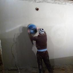 Tomas-Bud Tomasz Urbańczyk - Tynkarz szlifuje ścianę za pomocą szlifierki oscylacyjnej. Widoczne przewody elektryczne i nierówności na podłodze. Realizacja tynkowania ścian.