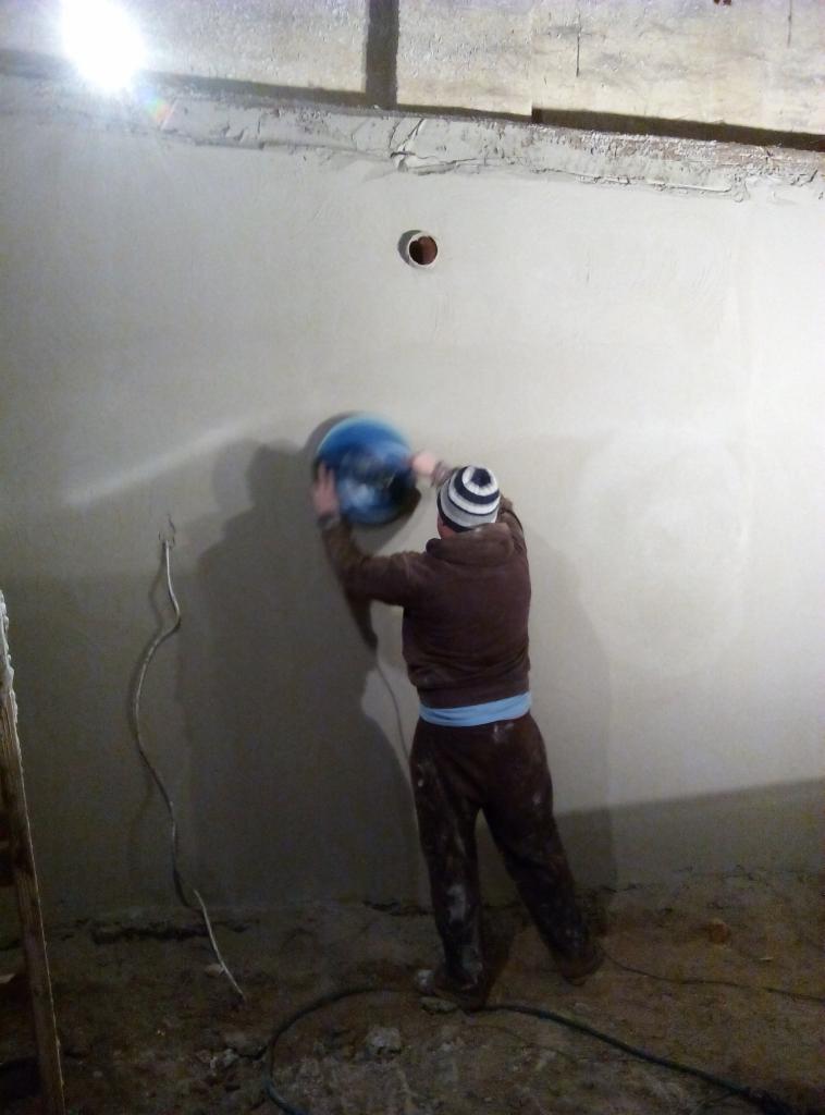 Tynkarz szlifuje ścianę za pomocą szlifierki oscylacyjnej. Widoczne przewody elektryczne i nierówności na podłodze. Realizacja tynkowania ścian.