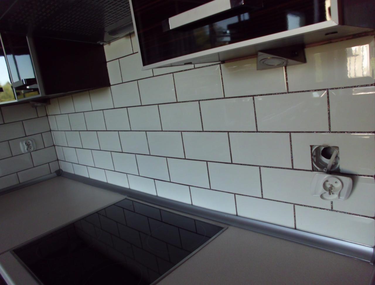 Białe, prostokątne płytki z ciemną fugą na ścianie w kuchni, widoczny fragment blatu i okapu. Gniazdko elektryczne i otwór na puszkę instalacyjną.