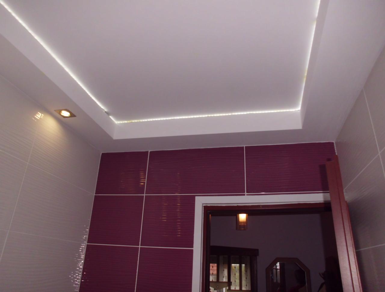 Nowoczesna łazienka z podwieszanym sufitem i oświetleniem LED. Bordowe płytki na ścianie kontrastują z białym sufitem i jasnymi płytkami. Widoczny fragment futryny drzwi.