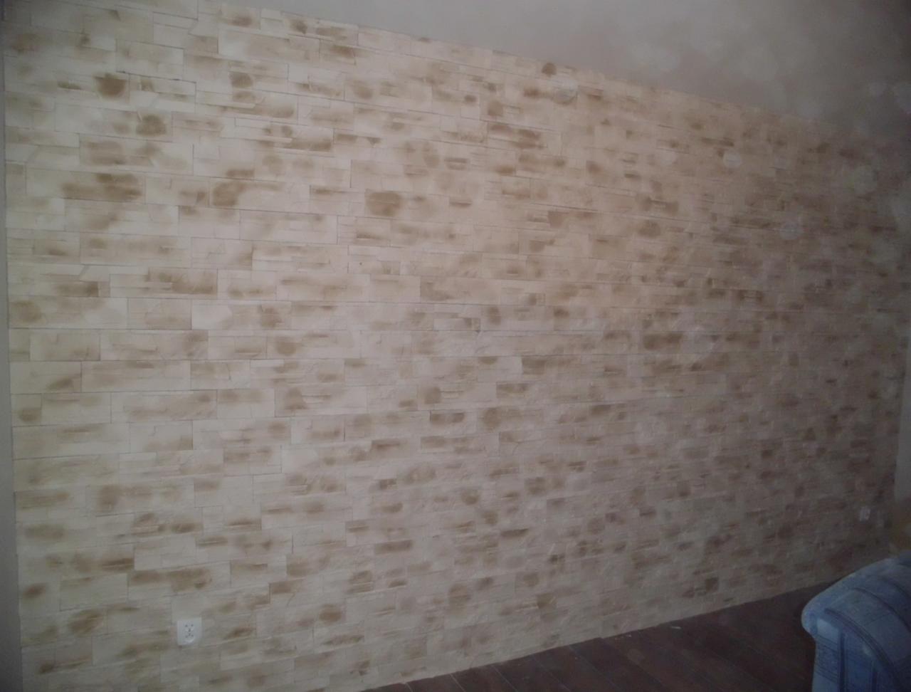 Ściana z dekoracyjną okładziną imitującą cegłę w odcieniach beżu i brązu, z widocznym gniazdkiem elektrycznym u dołu i fragmentem podłogi z paneli.