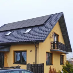 Żółty dom jednorodzinny z ciemną dachówką i panelami słonecznymi na dachu, widoczne okna dachowe i balkon z czarną balustradą.