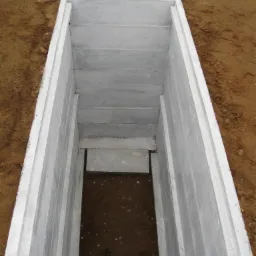 Otwarty prefabrykowany element betonowy, prawdopodobnie piwnica cmentarna, umieszczony w wykopie ziemnym, widok z g&oacute;ry.