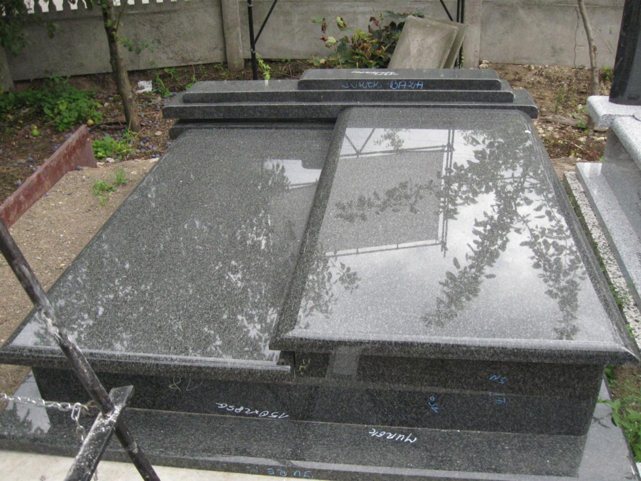 Elementy nagrobka dwuosobowego z ciemnego granitu, z widocznymi napisami i odbiciem drzewa na polerowanej powierzchni, składowane na zewnątrz.