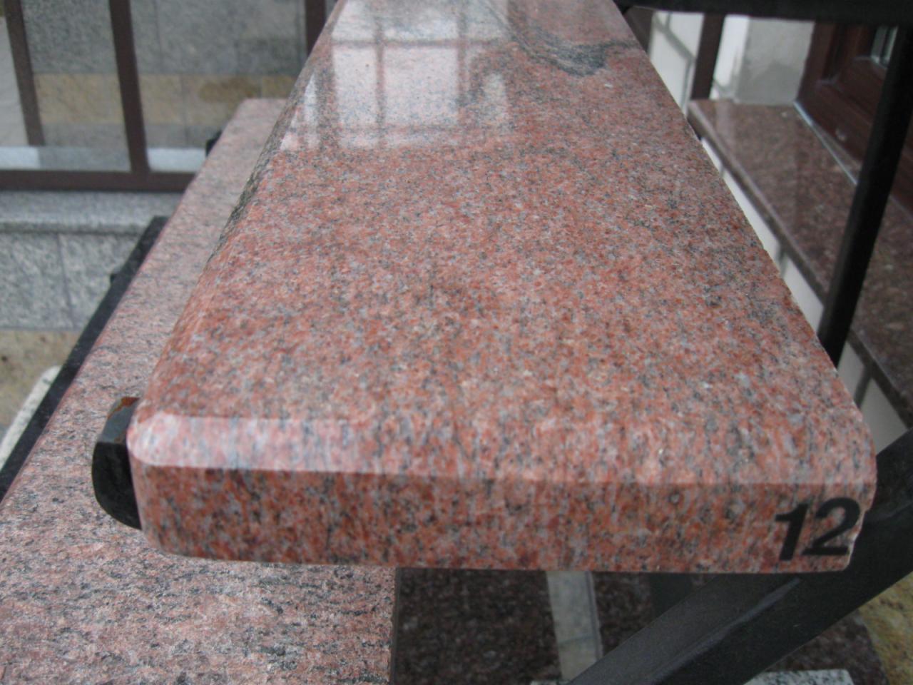 Czerwono-szary blat granitowy z zaokrąglonym brzegiem i numerem '12', zamontowany na czarnej metalowej konstrukcji, widoczne elementy balustrady i fragment okna w tle.