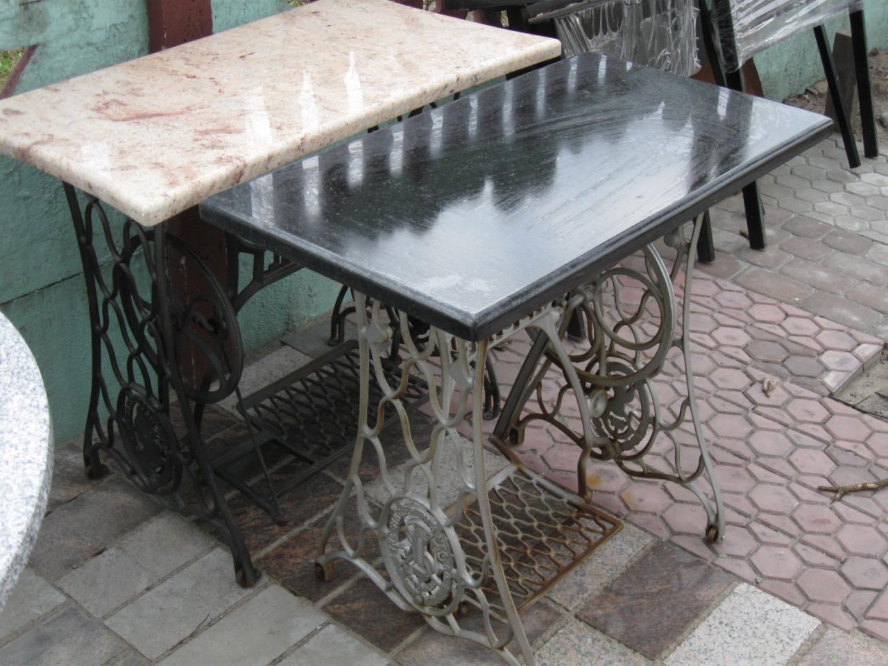 Dwa stoliki ogrodowe z blatami z jasnego i ciemnego granitu, osadzone na ozdobnych, metalowych nogach od starych maszyn do szycia, ustawione na kostce brukowej.