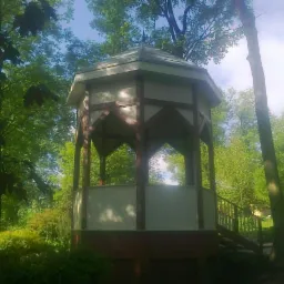 Ośmioboczna altana ogrodowa z drewnianą konstrukcją szkieletową i białymi panelami, otoczona zielenią drzew i krzewów w słoneczny dzień.