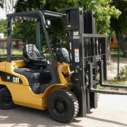 Żółty wózek widłowy marki CAT o udźwigu 2.5 tony, zaparkowany na zewnątrz, z placem zabaw dla dzieci widocznym w tle za ogrodzeniem.